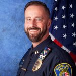 a uniformed police officer from the Camillus Police Department. The officer is wearing a dark blue police uniform adorned with various insignia and badges. On the left side of the chest, there are two rectangular nameplates and a badge with an eagle emblem. Above these, there are several service ribbons arranged in rows. The shoulder patch on the right arm reads "CAMILLUS POLICE" in yellow letters, with "PROFESSIONALISM INTEGRITY" written below it, encircling an American flag emblem. In the background, there is an American flag positioned to the right of the officer against a blue textured backdrop.