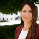 Individual with long braided hair in maroon blazer and white shirt stands outdoors; background shows greenery and a white building