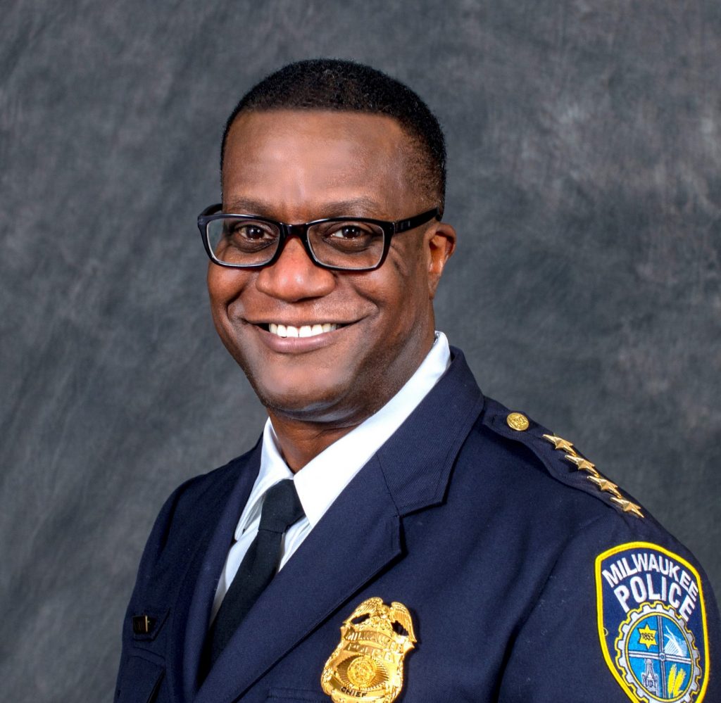 Person wearing a dark Milwaukee Police dress uniform with gold buttons, shoulder braid, badge, and embroidered patch, posed against a gray textured backdrop