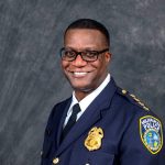 Milwaukee Police officer in dark navy dress uniform with gold badge, shoulder stars, and department patch, standing against a textured gray backdrop