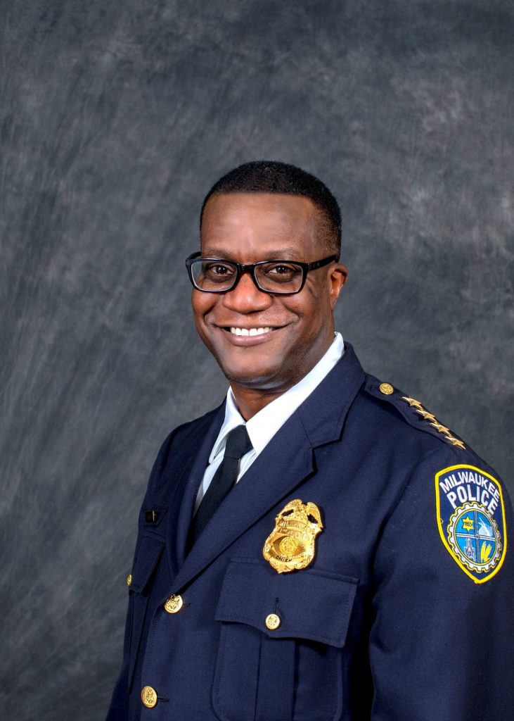Milwaukee Police officer in dark navy dress uniform with gold badge, shoulder stars, and department patch, standing against a textured gray backdrop