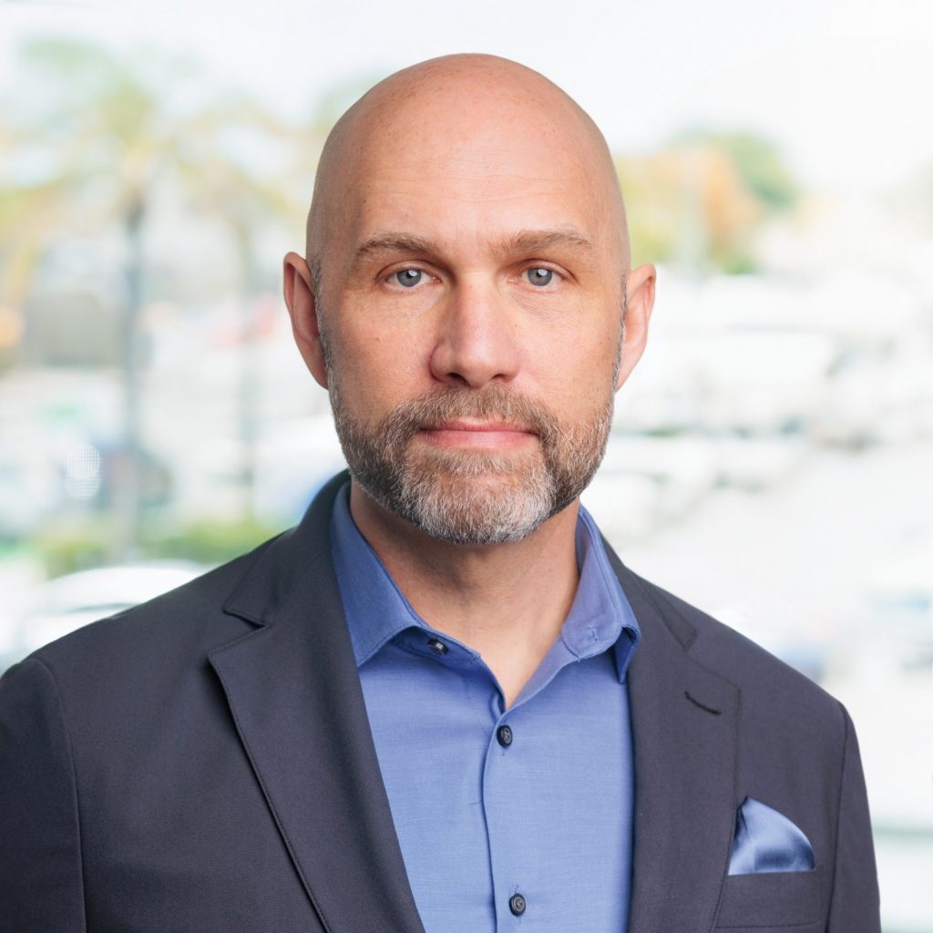 Individual wearing a dark blazer over a light blue dress shirt with matching pocket square stands before a softly blurred office setting with windows, palm trees, buildings, and daylight visible in the background