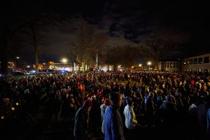 Large nighttime crowd gathered in a park during a candlelight vigil, illuminated by streetlights and handheld candles, with trees, nearby buildings, and emergency vehicle lights visible around the perimeter.