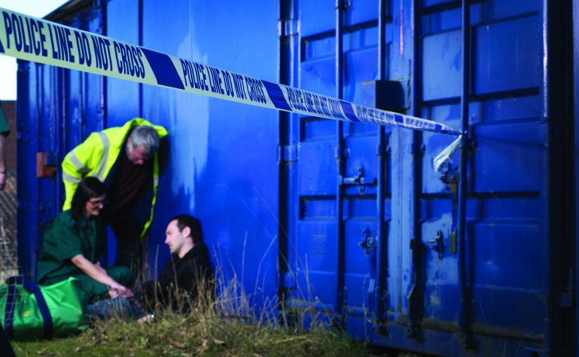 Police line tape marks crime scene beside blue shipping container; two responders, one in high-vis gear, assist person lying on the ground