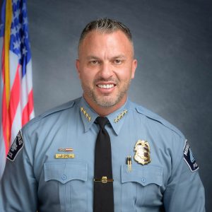 Police officer in light blue uniform shirt with shoulder patches, black tie, and multiple badges and pins, standing before an American flag backdrop