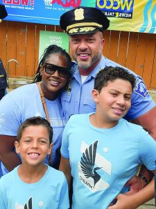 Uniformed officer stands with family of four; two kids wear blue shirts with bird logo; WCCO News Talk banner visible in background; outdoor setting
