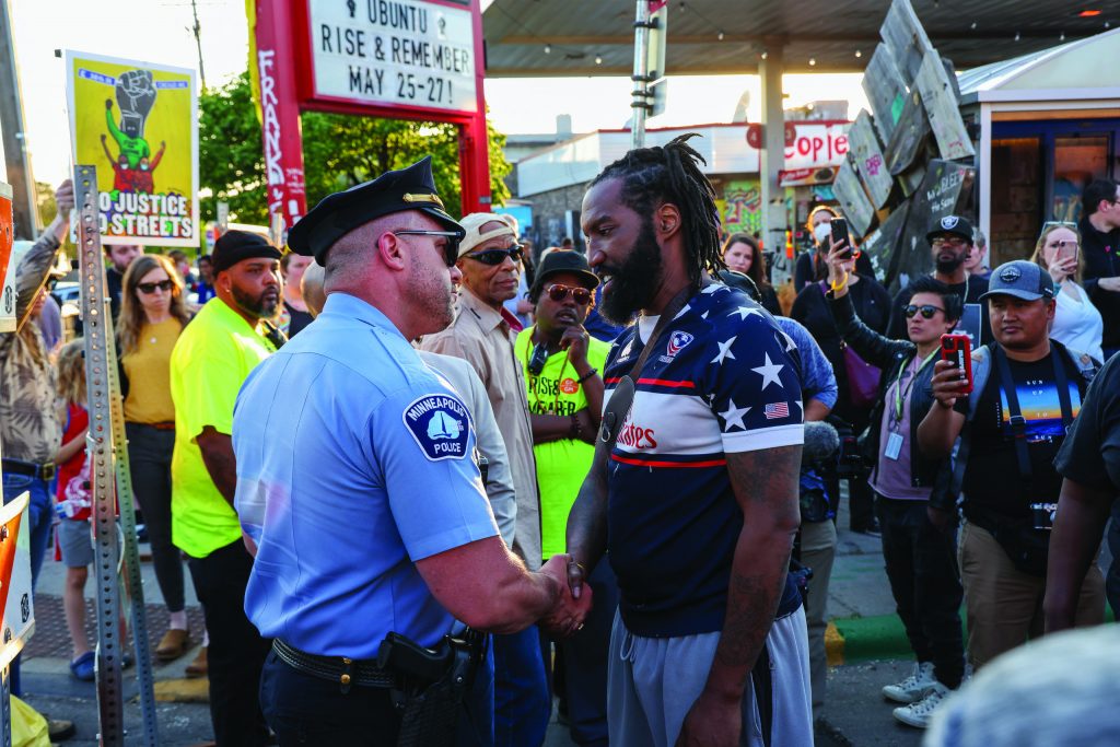 Minneapolis officer in uniform shakes hands with man in star-spangled shirt; outdoor event with signs, crowd, and festive atmosphere.