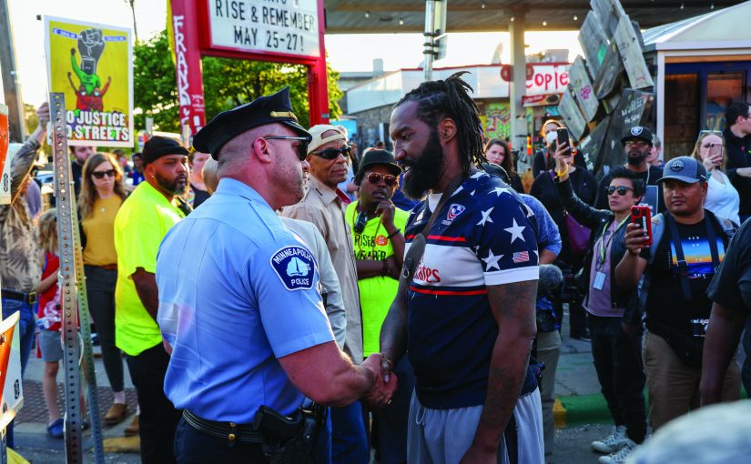 Minneapolis officer in uniform shakes hands with man in star-spangled shirt; outdoor event with signs, crowd, and festive atmosphere.
