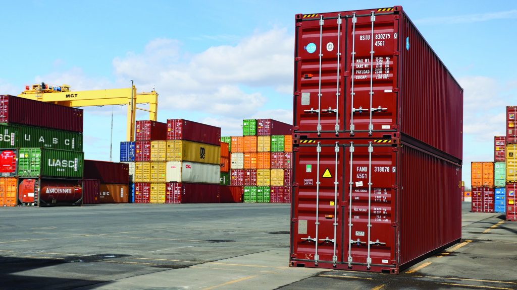 Shipping yard with stacked red, green, yellow, orange containers; yellow crane labeled 'MGT' in background under partly cloudy sky