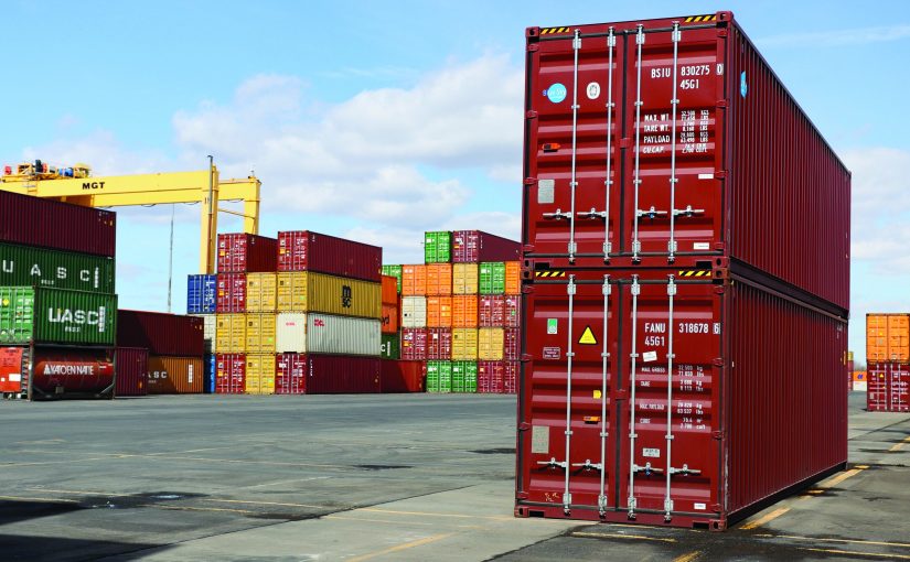 Shipping yard with stacked red, green, yellow, orange containers; yellow crane labeled 'MGT' in background under partly cloudy sky