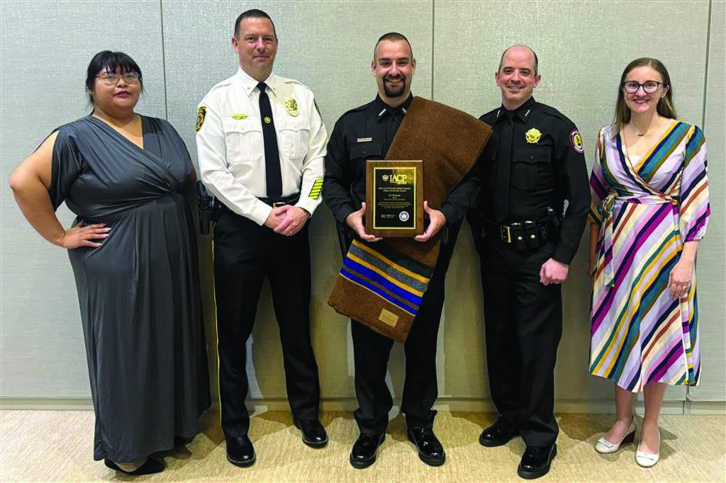Five people stand side by side indoors against a neutral wall, including uniformed officers and civilians, with one person holding a framed IACP award plaque and ceremonial textile
