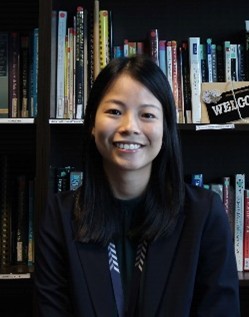 Person in a dark blazer and patterned lanyard sits before tall bookshelves filled with assorted books, including a small “WELCOME” sign on the right shelf.