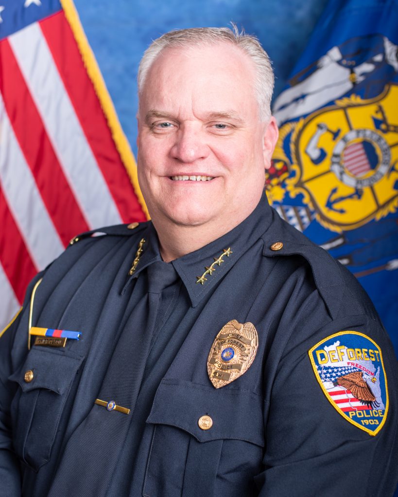 Uniformed police chief in dark dress uniform with gold collar stars, badge, service ribbon, tie, brass buttons, and DeForest Police shoulder patch poses before U.S. and Wisconsin state flags against a blue studio backdrop