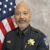 Officer in uniform with badge, nameplate, and shoulder patch stands before U.S. flag; formal portrait-style setting