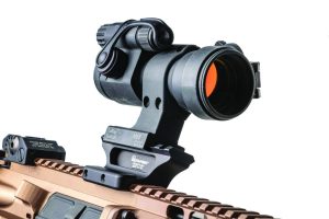 Close-up of a black M68 CCO optic mounted on a tan rifle rail, showing adjustment knobs, protective caps, and sturdy LaRue Tactical mount against a white background