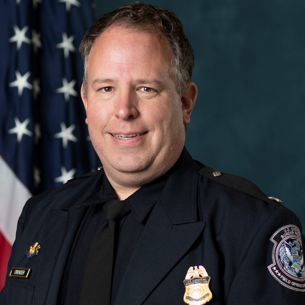Law enforcement officer in uniform with badge, name tag “Orender,” and emblem shoulder patch, standing before an American flag backdrop