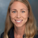 Person with long wavy blonde hair wearing a dark collared shirt and small hoop earrings, posed against a smooth blue-gray studio background
