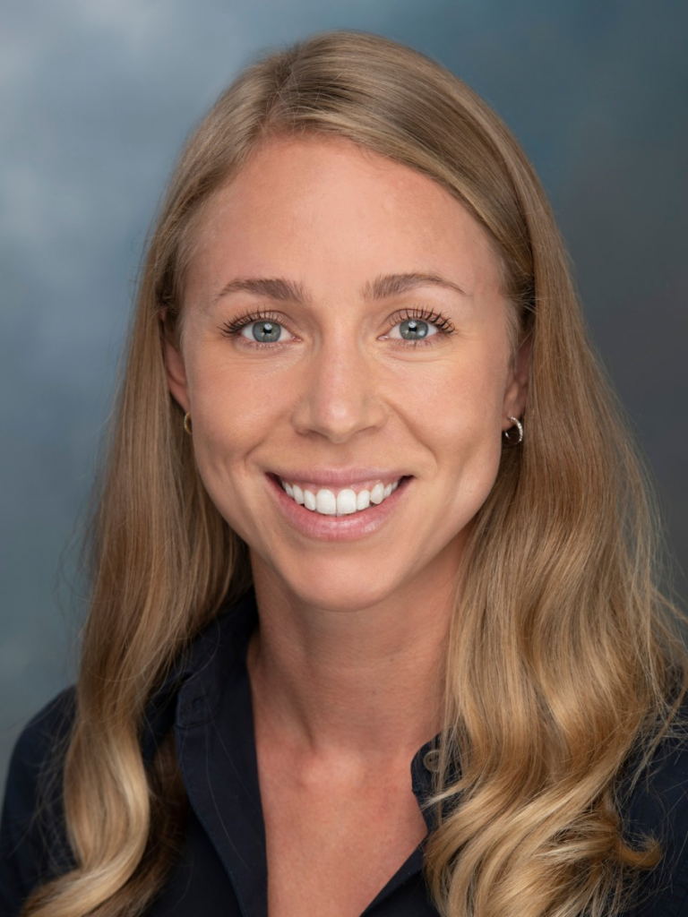 Person with long wavy blonde hair wearing a dark collared shirt and small hoop earrings, posed against a smooth blue-gray studio background