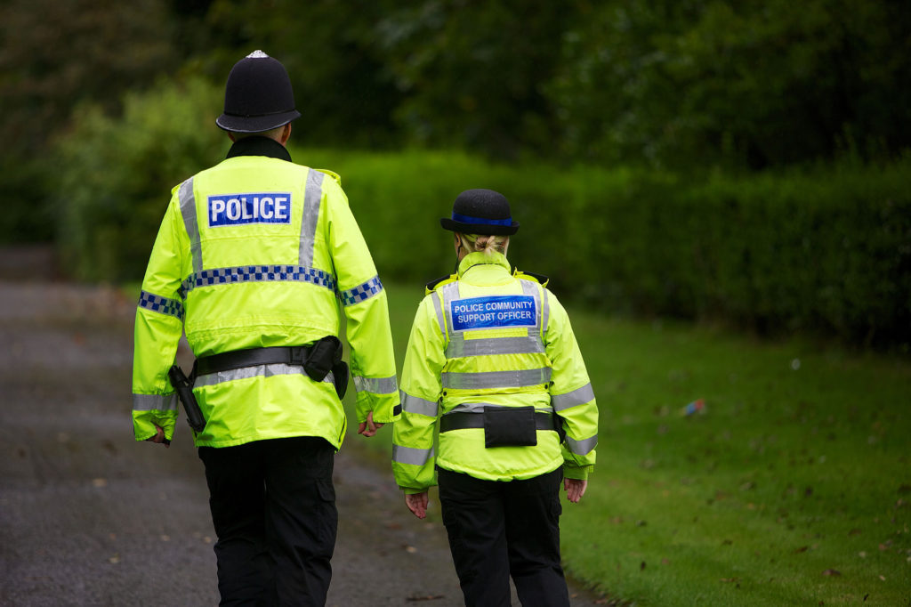 Two officers in high-vis jackets labeled 'POLICE' and 'POLICE COMMUNITY SUPPORT OFFICER' walk along a path bordered by greenery