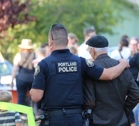 Portland Police officer in dark uniform with visible shoulder patches and duty belt walks outdoors with arm around an older person in a suit and flat cap during a community event.