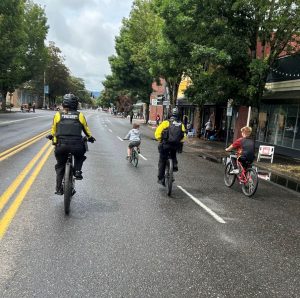 Two Portland Police officers in yellow and black bike uniforms ride bicycles alongside two children on a wet city street lined with trees, storefronts, and scattered spectators.