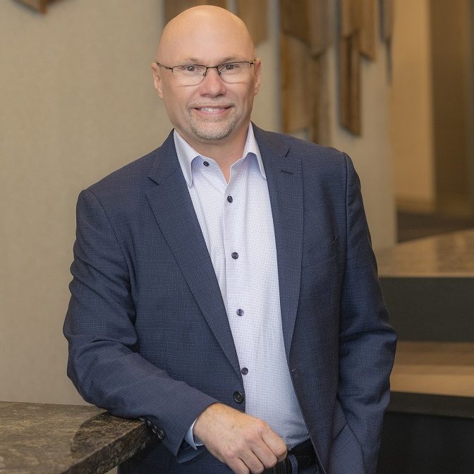 Person in a dark blue suit with light patterned dress shirt, standing indoors near a stone countertop with geometric wood wall panels in background