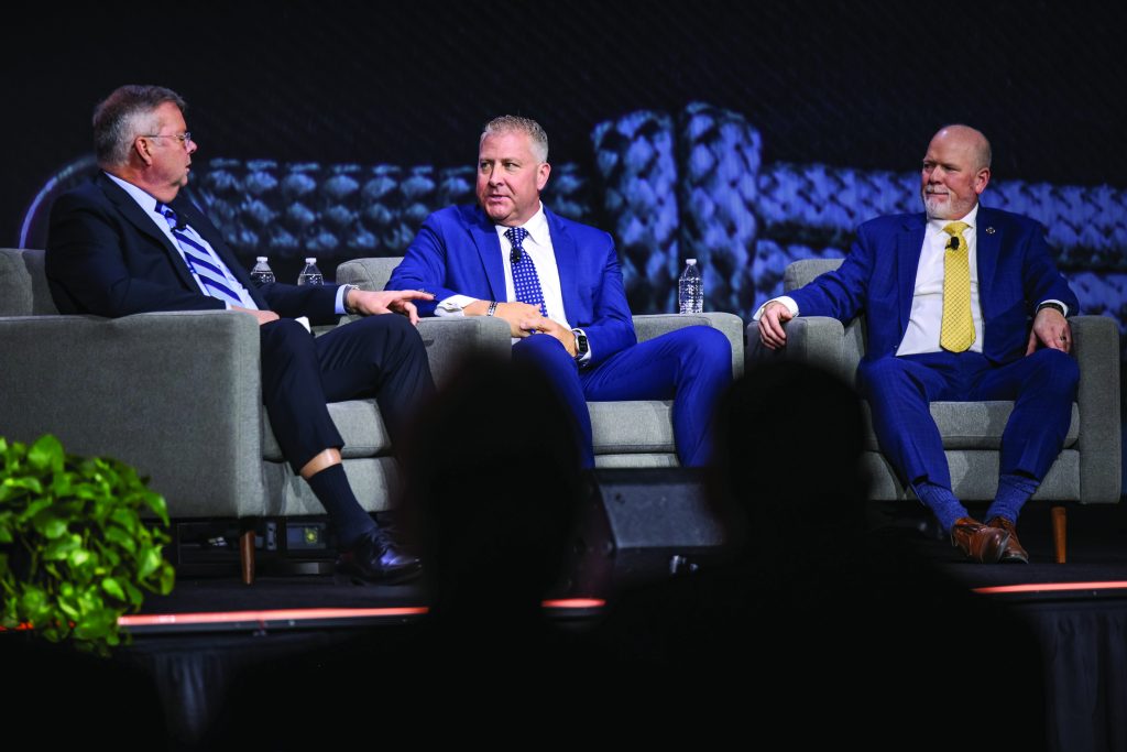 Three individuals in suits sit on stage in gray armchairs during a panel discussion, with microphones, water bottles, and a textured backdrop behind them