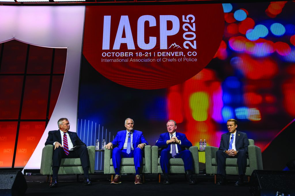Four individuals in suits seated on stage in gray armchairs during an IACP 2025 panel discussion, with large event signage and colorful lights in the background