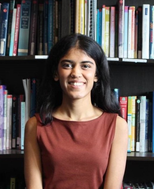 Individual wearing a sleeveless rust-colored top sits before tall bookshelves filled with books, journals, and reference volumes, with dark shelving, labels, and library furnishings visible throughout the background