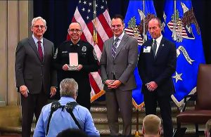 Four individuals stand before American and blue flags; one in police uniform holds award while photographer captures moment from side angle