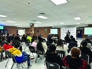 Audience seated in rows faces speaker in room with brick walls, ceiling tiles, and two screens showing presentations on either side of the front