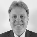 Grayscale image of person in suit with striped tie and dual-flag lapel pin; face blurred to emphasize formal attire and symbolic accessory.