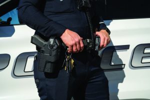 Close-up of an officer’s hands resting near a duty belt holding a holstered tool, keys, and radio gear while standing beside a marked patrol vehicle in daylight