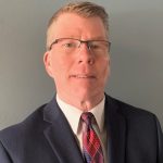 Individual in dark blue suit, white shirt, red-blue patterned tie stands before plain gray background