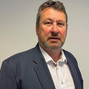 Individual in dark blue suit jacket and white shirt with gold buttons stands against plain light background;