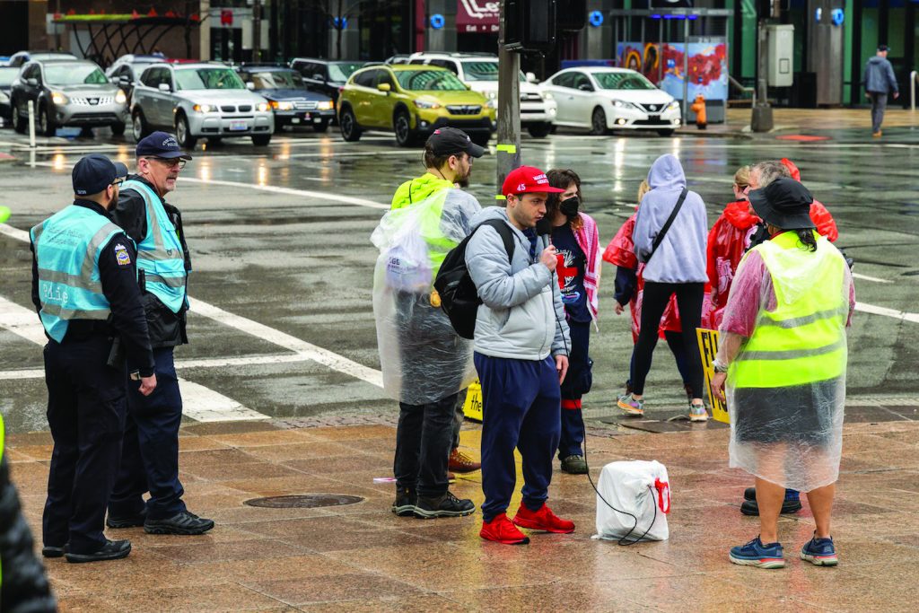 Columbus police dialogue team officers in reflective vests walk alongside protesters carrying signs and flags near a fenced lawn and columned government building