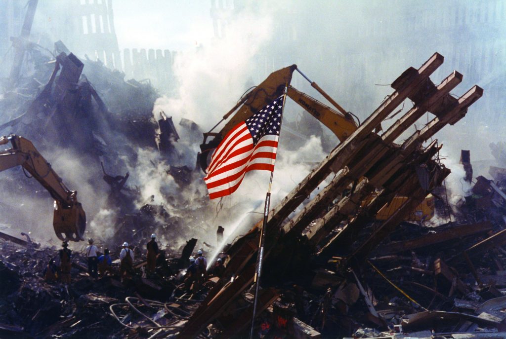 American flag stands in rubble of collapsed building; smoke rises as construction machinery and workers operate in background.