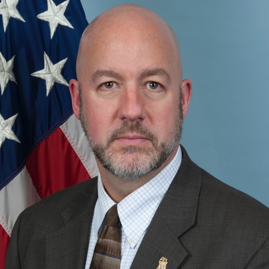 An individual in a suit jacket, collared shirt, and striped tie stands before a blue backdrop with a U.S. flag positioned to the left side of the portrait.