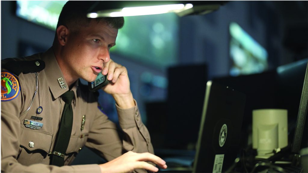 Uniformed officer speaks on phone while typing on laptop under desk lamp; office background includes shelves, papers, and equipment.