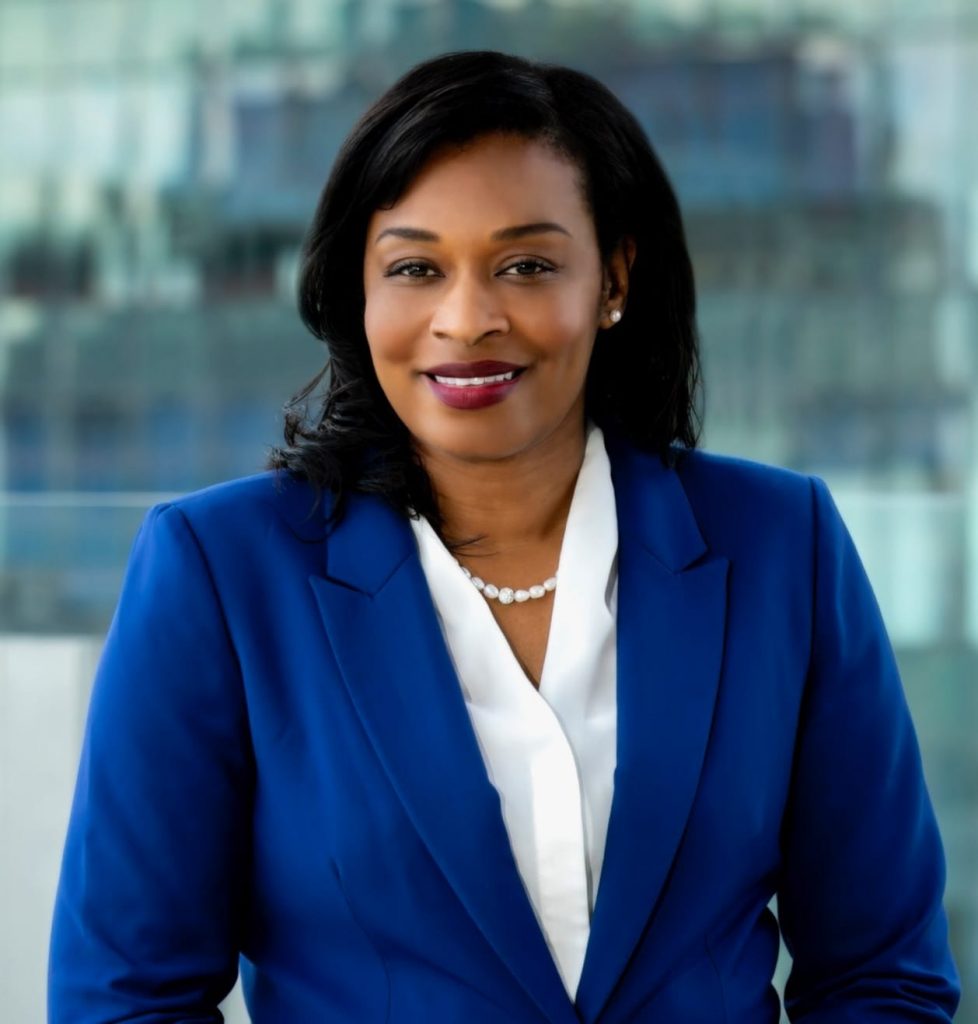 Professional portrait shows an individual in a blue blazer and white blouse with pearl necklace, standing before a blurred cityscape background with glass and skyline