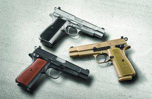 Three semi-automatic pistols on textured gray surface; finishes include silver with black grip, black with brown grip, and tan with matching grip