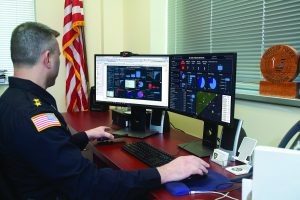 Uniformed officer at desk with dual monitors showing data dashboards; American flag and 'Chiefs of Police Association' plaque on windowsill behind