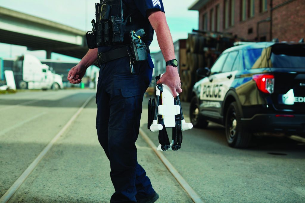 Police officer in tactical gear holding a folded drone, standing near a patrol vehicle in an industrial area.