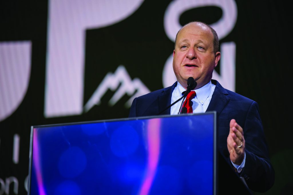 Individual in a dark suit and red tie speaks at a podium with dual microphones on stage, large event signage and abstract graphics visible in the background