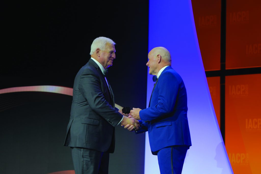 Two individuals in formal suits shaking hands on a stage with curved backdrop and IACP logo panels, under bright event lighting during a ceremonial exchange