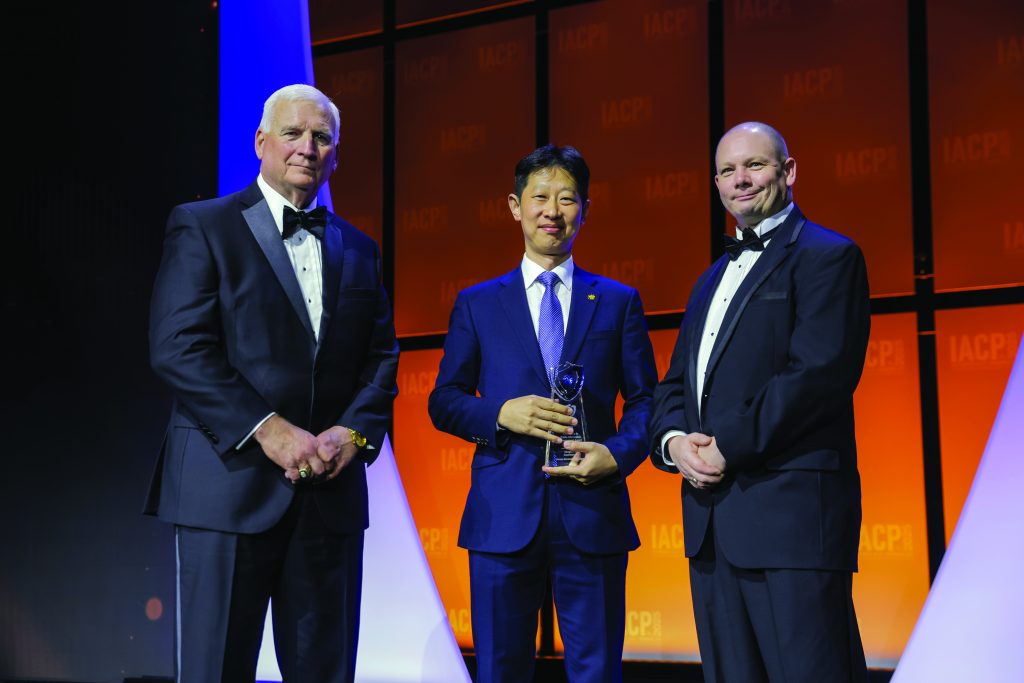 Three individuals in formal suits stand on stage with orange IACP backdrop; center person holds a glass award while others pose under bright event lighting.
