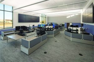 Modern public safety operations center with multiple workstations, large wall-mounted screens, gray desks, ergonomic chairs, and floor-to-ceiling windows
