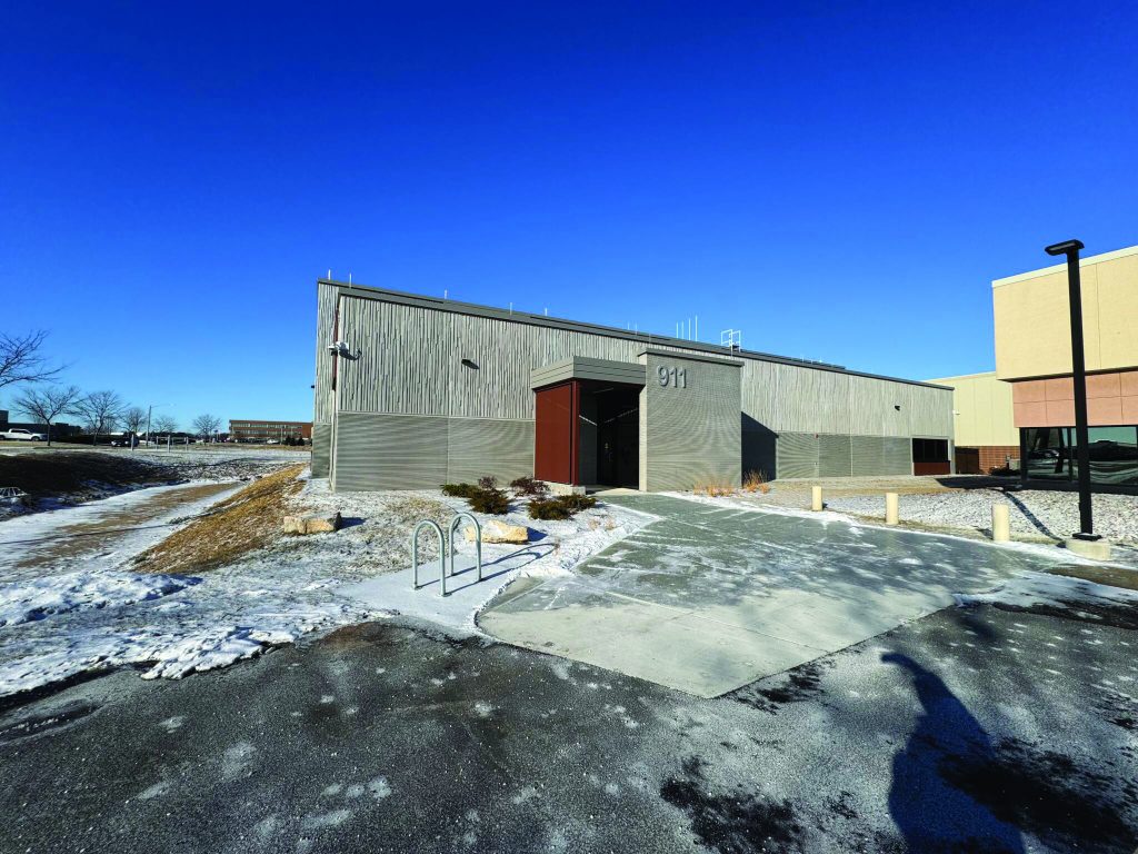 Exterior view of a modern public safety facility with gray metal siding, red entrance door, bike rack, snow-covered ground, and clear blue sky in background