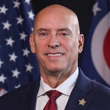Individual in a dark suit jacket, white shirt, and red patterned tie with star‑shaped lapel pin stands before U.S. and Ohio state flags on a formal backdrop
