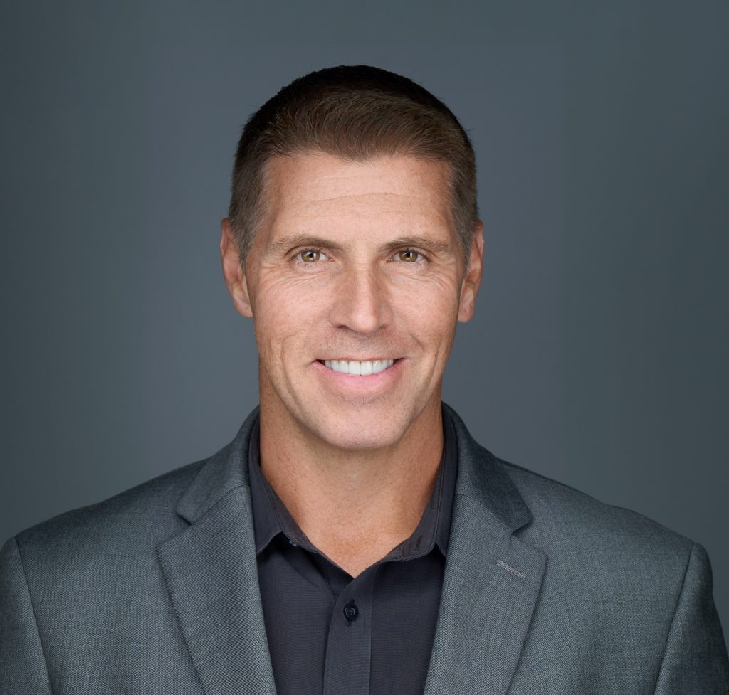 Studio portrait of an individual wearing a gray textured blazer over a dark collared shirt against a smooth gray backdrop with professional lighting